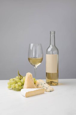 Different types of cheese, grapes, bottle and white wine glass on gray clipart