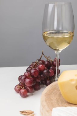 Partial view of maasdam cheese on wooden board, wine glass, almond and grapes on gray clipart