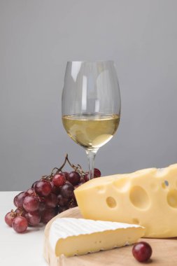 Brie and maasdam cheese on wooden board, white wine glass and grapes on gray clipart