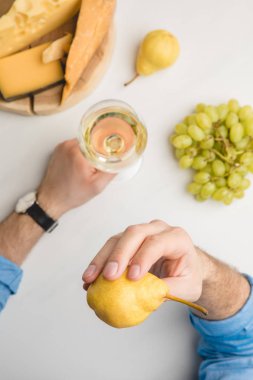 Cropped image of man with pear and wine glass, grapes and different types of cheese on wooden board on white clipart