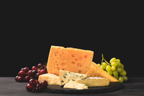 Different types of cheese on board with grapes on black 