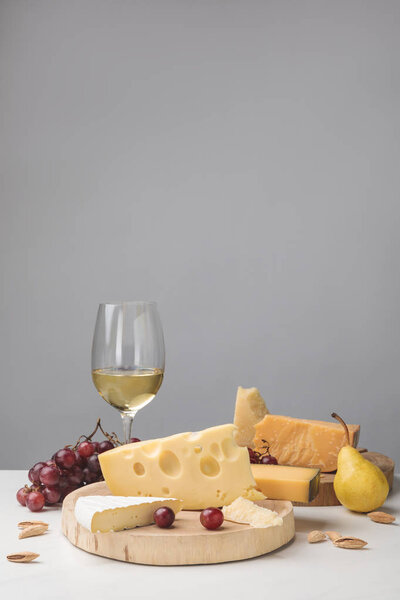 Different types of cheese on wooden boards, wine glass, fruits and almond on gray