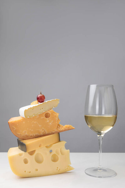 Close up view of white wine glass and grape on stack of different types of cheese on gray