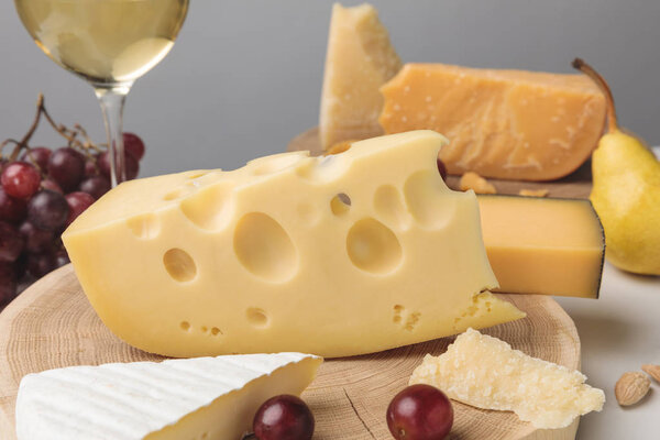 Closeup shot of different types of cheese on wooden board, pear, grapes and wine glass on gray
