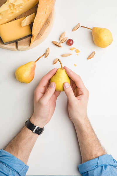 Cropped image of man with pear, almond and different types of cheese on wooden board on white