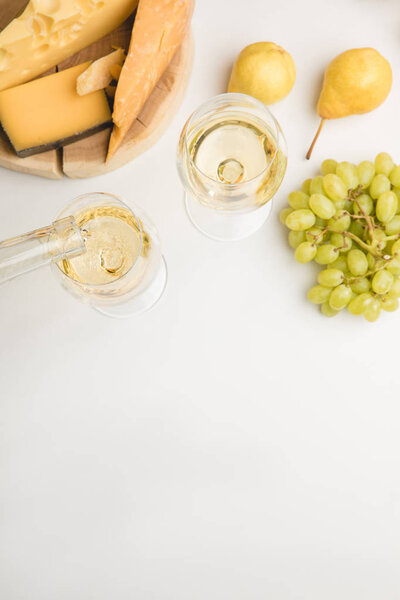 Top view of wine pouring into glass, different types of cheese on wooden board, grapes and pears on white