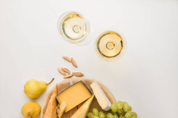 Top view of different types of cheese on wooden board, wine glasses, almond and fruits on white