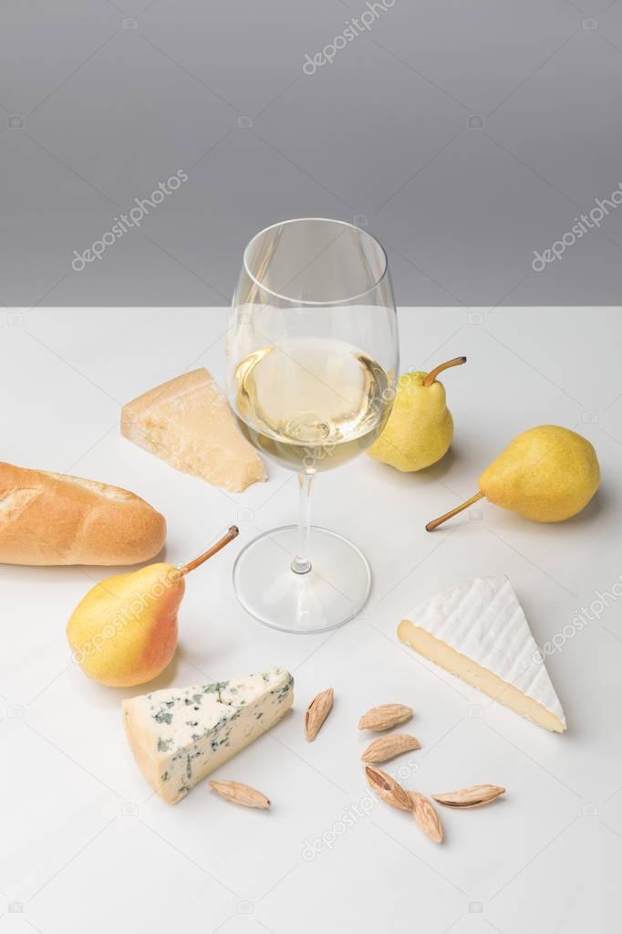 Elevated view of wine glass surrounded by pears, almond, baguette and different types of cheese on gray