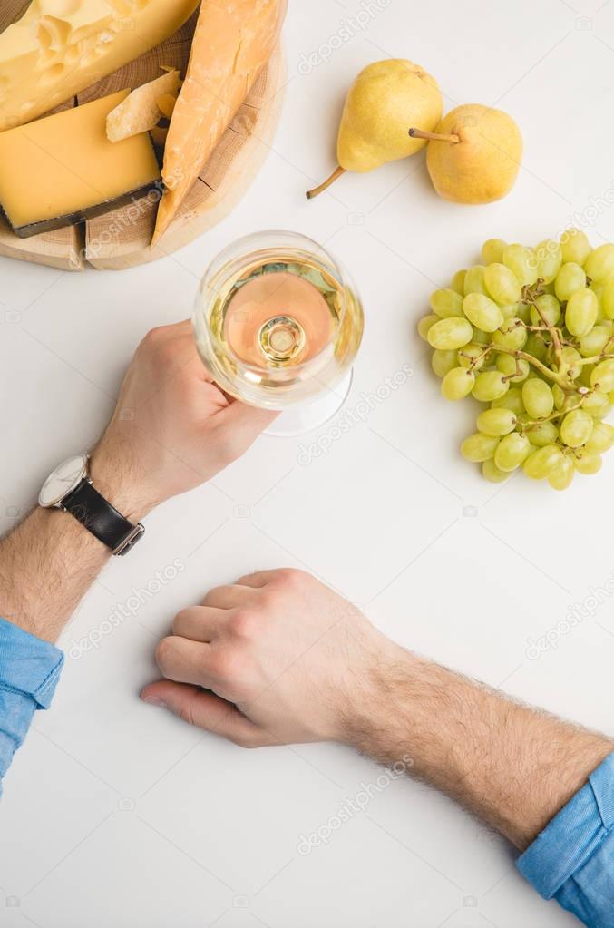 Cropped image of man with wine glass, different types of cheese on wooden board and fruits on white