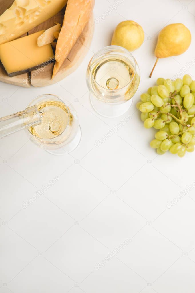 Top view of wine pouring into glass, different types of cheese on wooden board, grapes and pears on white