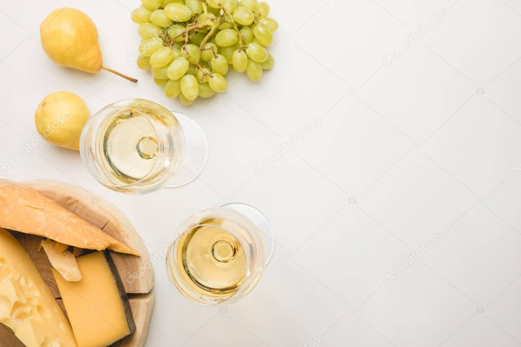 Top view of different types of cheese on wooden board, wine glasses and fruits on white