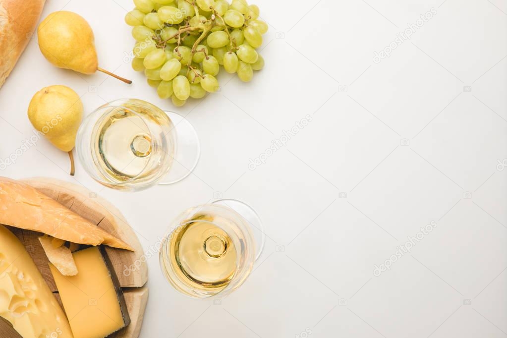 Top view of different types of cheese on wooden board, wineglasses and fruits on white