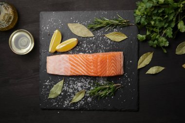 Piece of salmon with salt on black table with lemon and herbs clipart