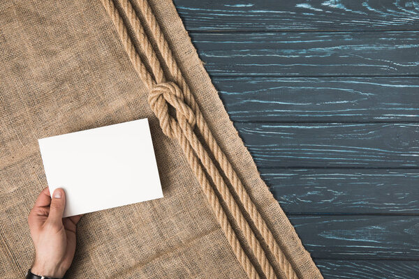 cropped image of man holding blank paper on sackcloth with knotted rope on wooden surface