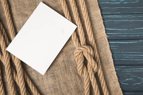 closeup shot of blank white paper on sackcloth and brown nautical knotted ropes on wooden tabletop