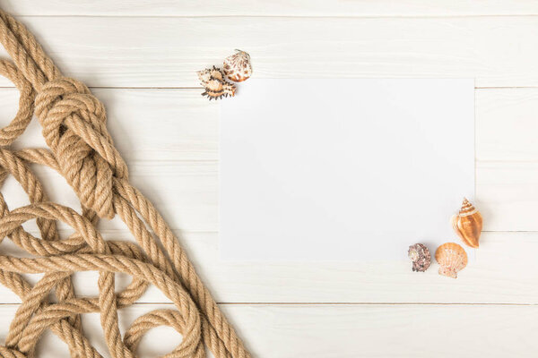 top view of brown nautical knotted rope and empty paper with seashells on white wooden surface