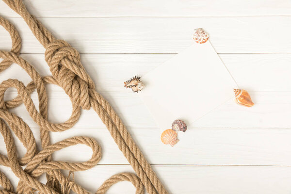 top view of brown nautical knotted rope and empty paper with seashells on white wooden surface