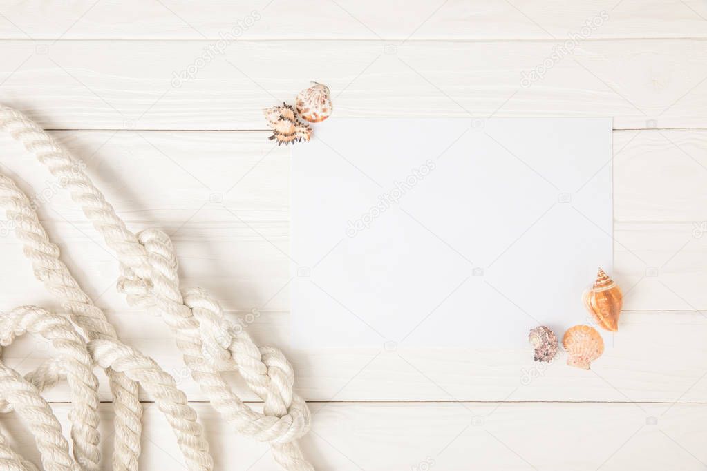 top view of white nautical knotted rope and empty paper with seashells on wooden surface