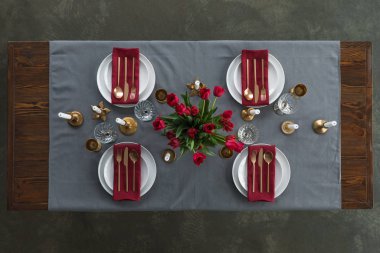 top view of rustic table setting with red tulips bouquet, tarnished cutlery, wine glasses, candles and empty plates on tabletop clipart