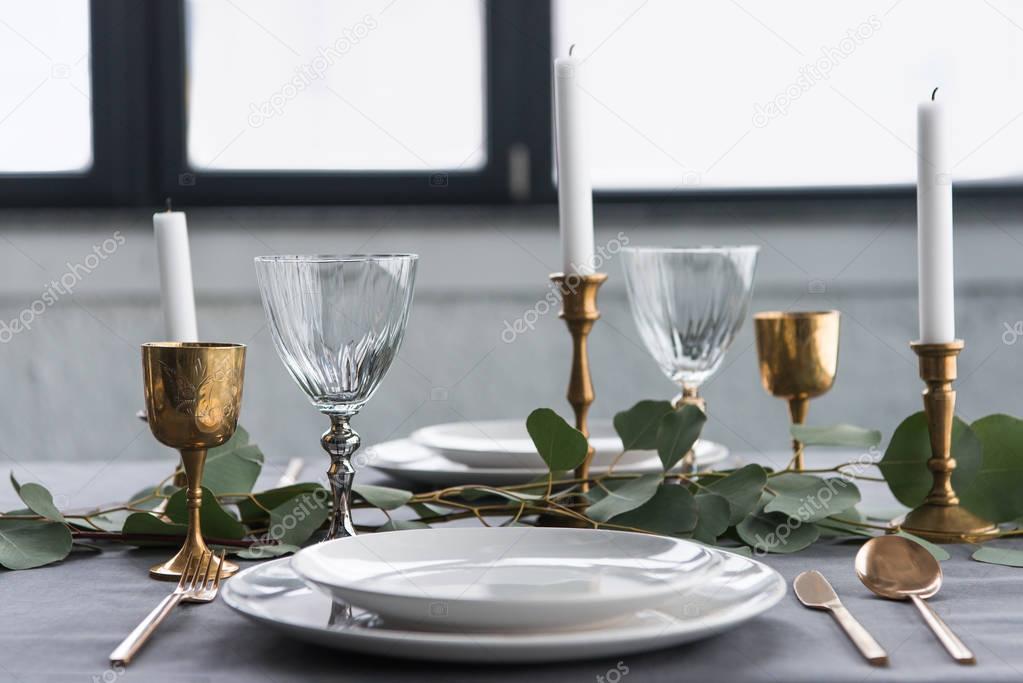 close up view of rustic table setting with wine glasses, eucalyptus, vintage cutlery, candles in candle holders and empty plates