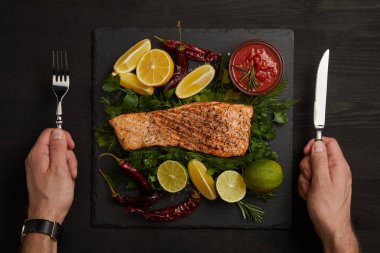 erkek kısmi görünümünü eller çatal bıçak takımı, ızgara somon steak, sos ve narenciye siyah yüzey üzerinde düzenlenmiş
