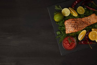 top view of grilled salmon steak, pieces of lime and lemon and sauce on black surface clipart