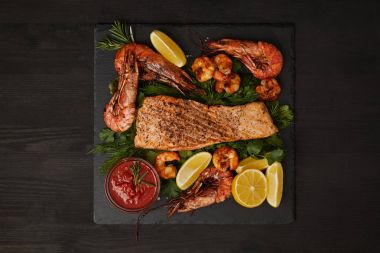 top view of grilled salmon steak, shrimps, pieces of lemon and sauce on black surface clipart