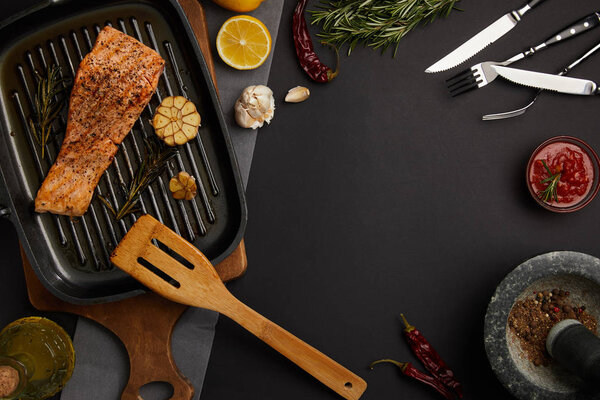 top view of arranged grilled salmon steak on wooden cutting board, ingredients, sauce and cutlery on black tabletop