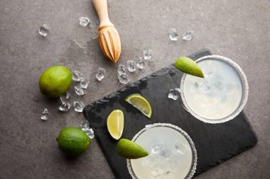 Top view of margarita cocktails with pieces of lime, ice cubes and wooden squeezer on grey tabletop clipart