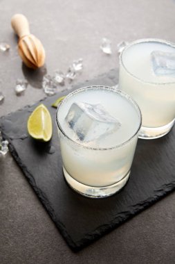 close up view of wooden squeezer, refreshing sour caipirinha cocktails with lime and ice on grey tabletop clipart