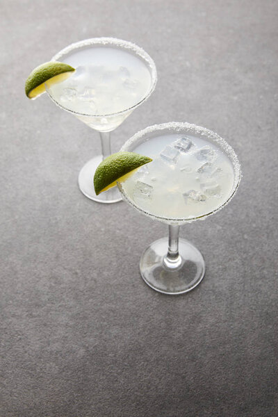 close up view of cold margarita cocktails with pieces of lime on grey tabletop