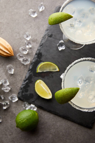 top view of alcohol margarita cocktails with pieces of lime, ice cubes and wooden squeezer on grey tabletop