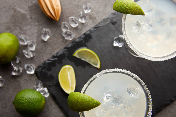 Top view of margarita cocktails with pieces of lime, ice cubes and wooden squeezer on grey tabletop