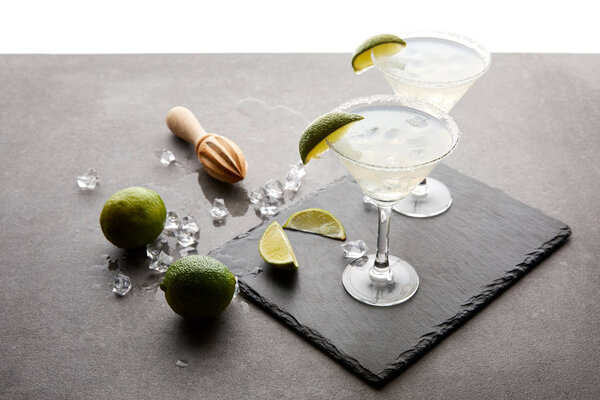 close up view of alcohol cocktails with pieces of lime, ice cubes and wooden squeezer on grey tabletop