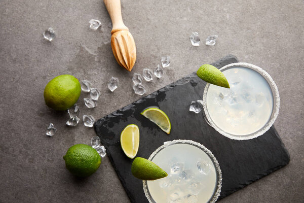 Top view of margarita cocktails with pieces of lime, ice cubes and wooden squeezer on grey tabletop