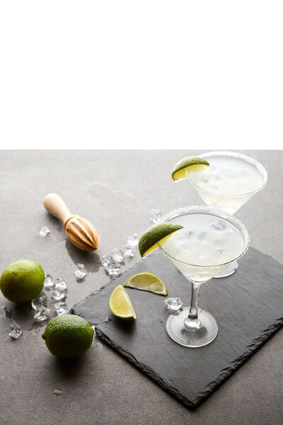 Close up view of margarita cocktails with lime pieces and wooden squeezer on grey surface  on white