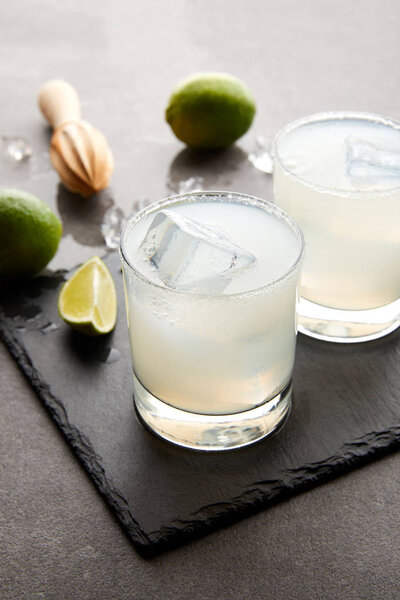 Close up view of wooden squeezer, refreshing margarita cocktails with lime and ice on grey tabletop