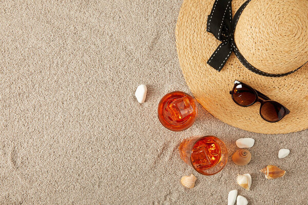 вид на соломенную шляпу, коктейли, ракушки и солнцезащитные очки на песке
