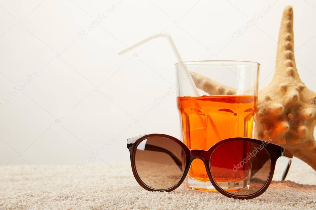 close up view of sunglasses, cocktail with straw and sea star on sand on grey backdrop