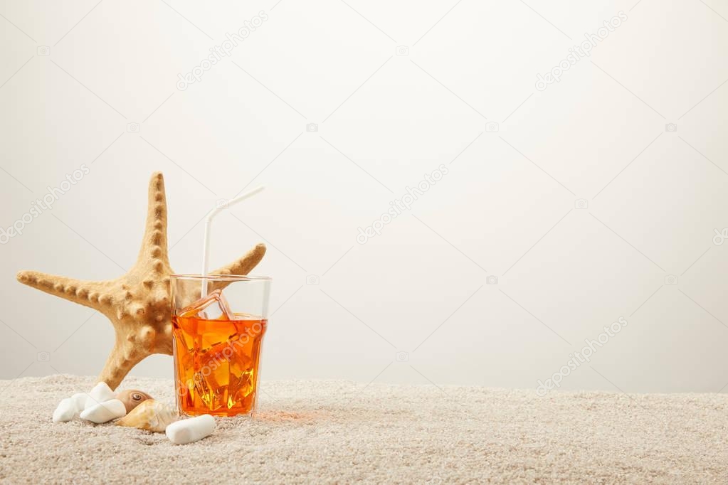 close up view of sea star, refreshing cocktail with straw and seashells on sand on grey background