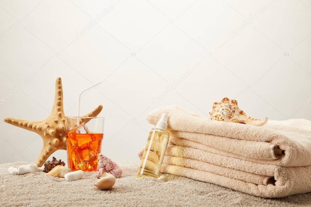 close up view of tanning oil, refreshing cocktail with ice, towels, sea star and seashells on sand on grey background