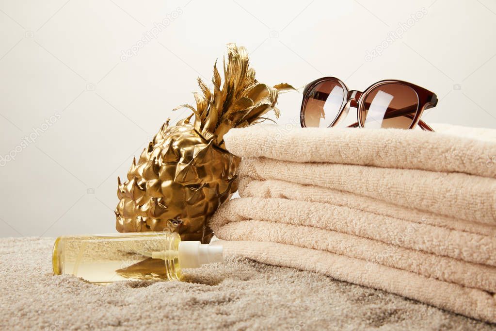 close up view of stack of towels, sunglasses, tanning oil and golden decorative pineapple on sand on grey backdrop
