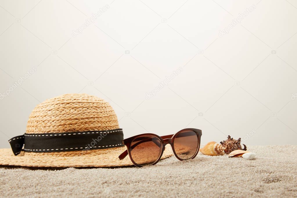 close up view of straw hat, sunglasses and seashells on sand on grey backdrop