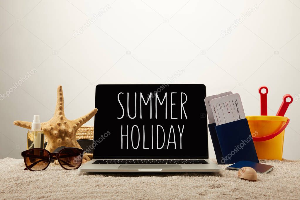 close up view of laptop with summer holiday lettering, sea star, sunglasses, passports with tickets and toy bucket on sand on grey background