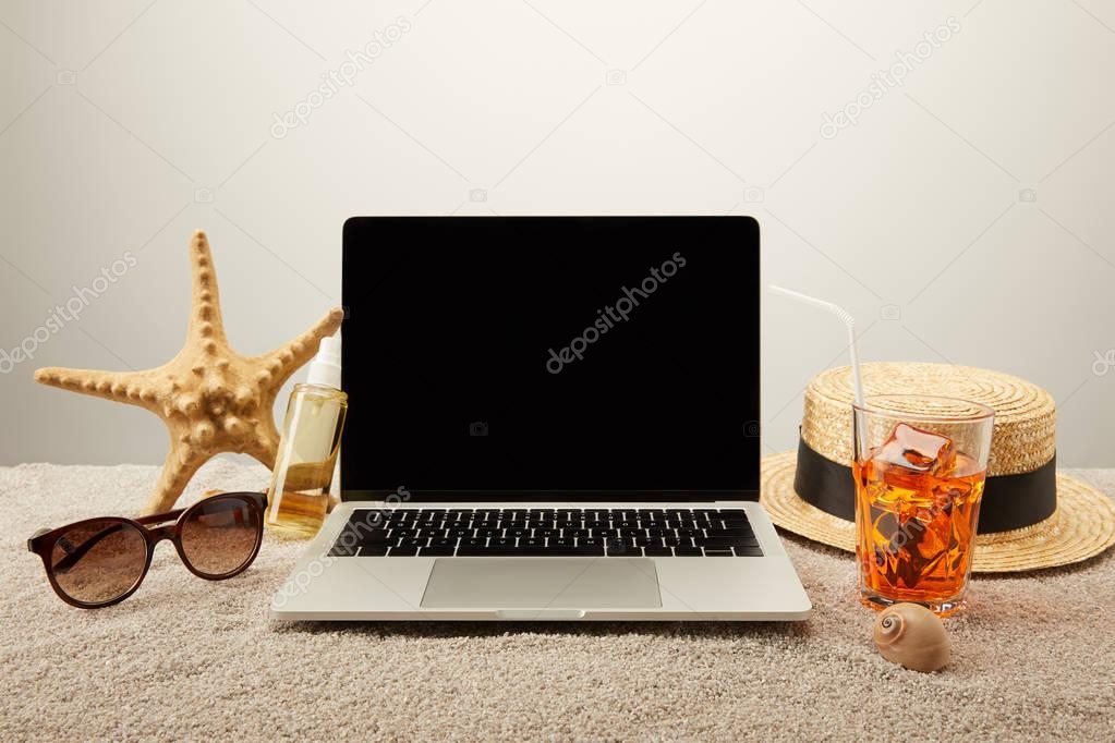 close up view of laptop with blank screen, refreshing cocktail and summer holiday accessories on sand on grey background