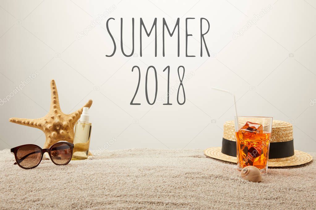 close up view of summer 2018 lettering, cocktail with ice, straw hat, sunglasses and tanning oil on sand on grey backdrop