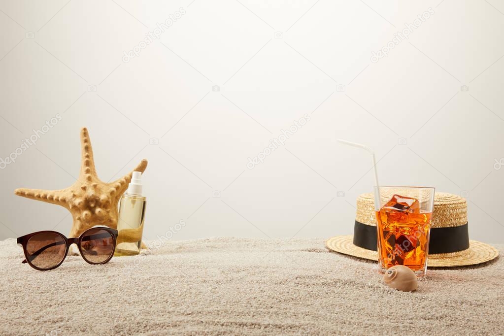 close up view of cocktail with ice, straw hat, sunglasses and tanning oil on sand on grey backdrop