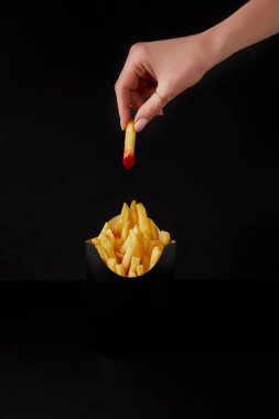 cropped shot of woman folding french fry poured into ketchup over box of fries isolated on black clipart