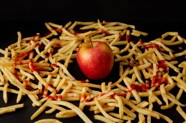red apple surrounded with french fries poured with ketchup isolated on black clipart