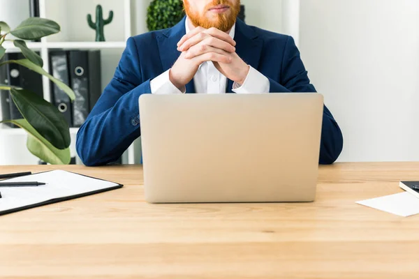 Partial view of bearded businessman in suit sitting at workplace with laptop in office — Stock Photo, Image